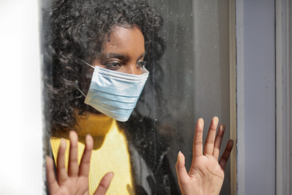 serious young ethnic lady in medical mask standing near window and looking away on street